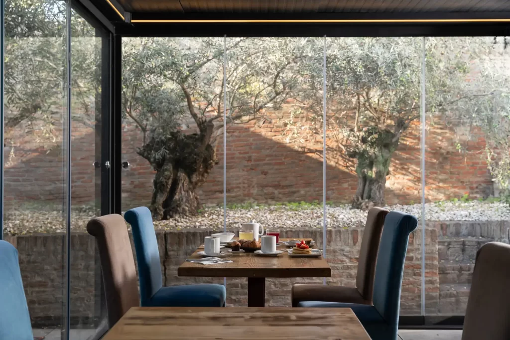 Tavolo apparecchiato in un ristorante luminoso con vista su un giardino con ulivi.