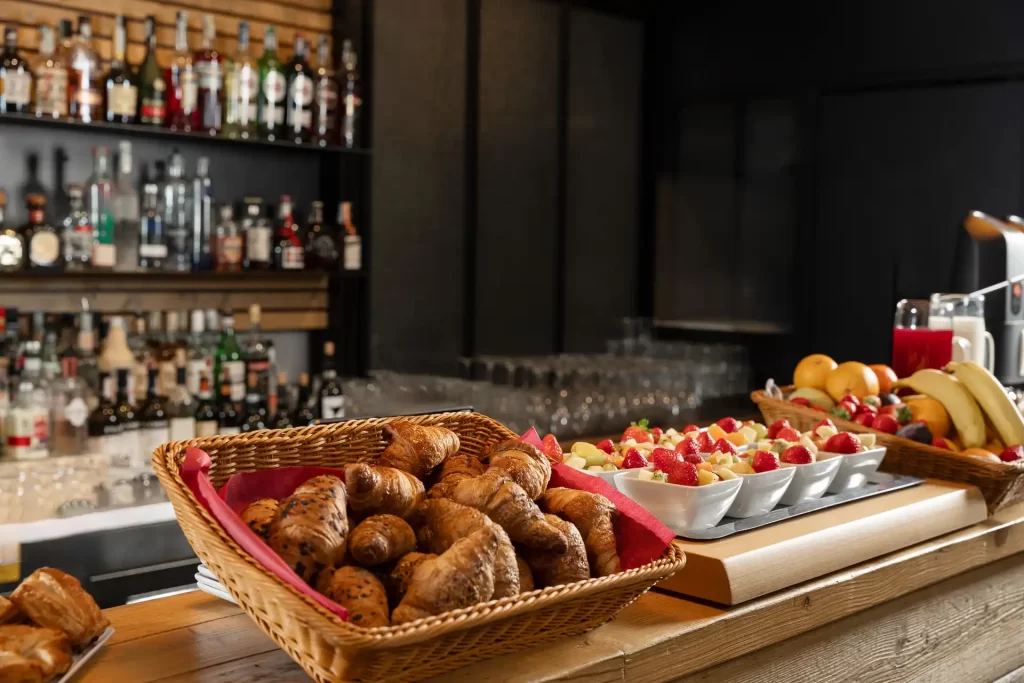 Cesto di croissant e frutta fresca su bancone di un bar con bottiglie sullo sfondo.