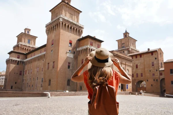Persona di spalle con zaino e cappello, osservando un grande castello storico in mattone.