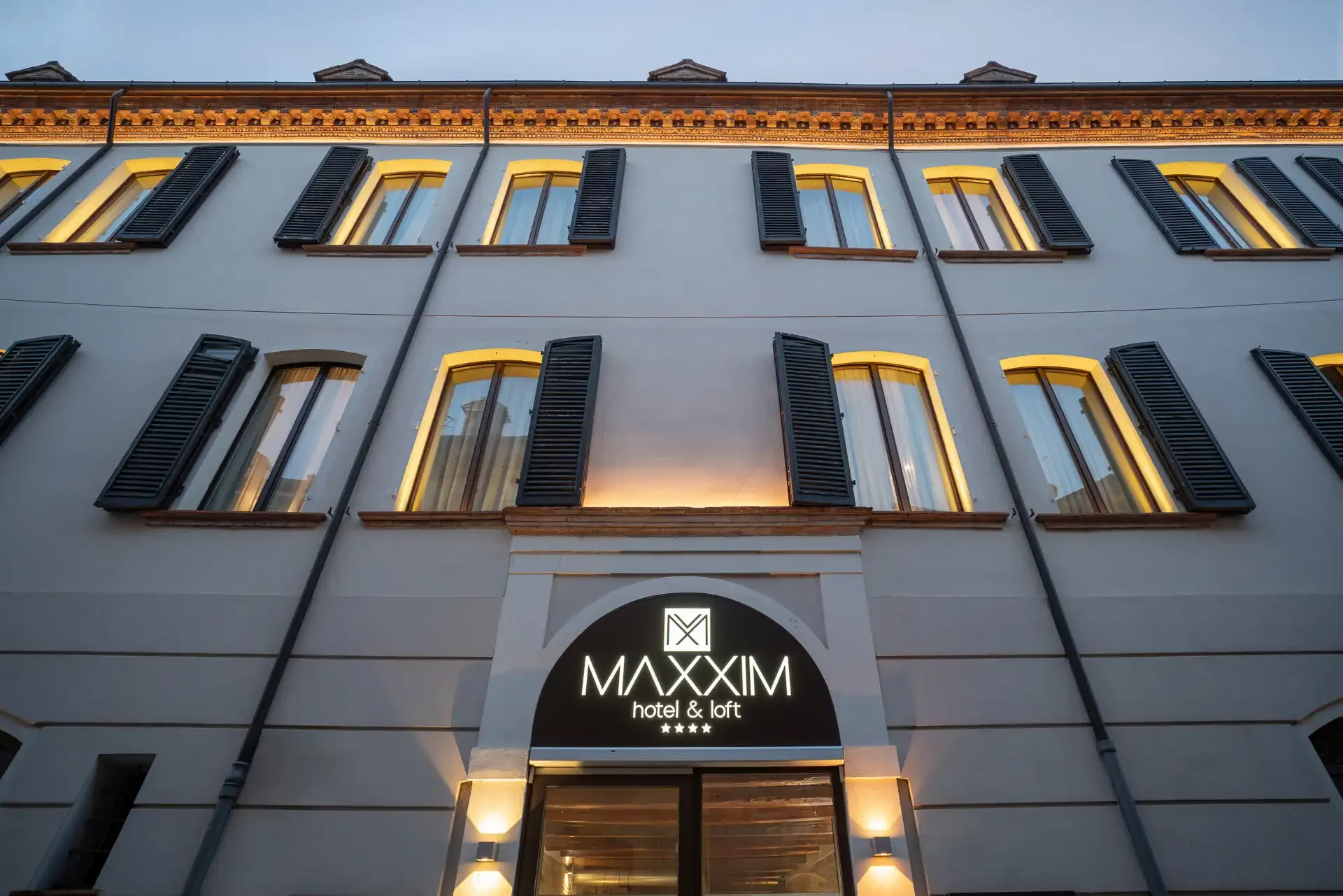 Facciata dell'hotel Maxxim con finestre illuminate e insegna al crepuscolo.